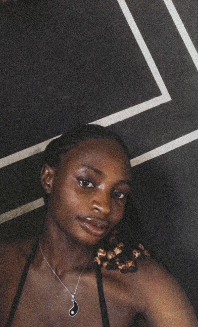 Selfie of a young woman with a calm expression, wearing a yin-yang pendant necklace. Her hair is braided and  adorned with beads, and she is posed against a dark painted wall with white lines