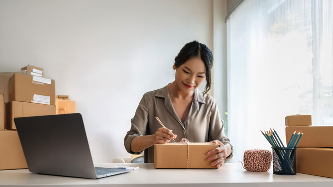 A small business owner packaging a product at a home-based workspace with shipping boxes, a laptop, and office supplies, symbolizing the fulfillment process of an e-commerce business.