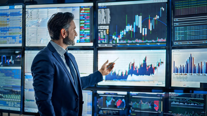 A financial analyst pointing at stock charts displayed on multiple large monitors, illustrating trends and data for major US stocks like Tyler Technologies (TYL), Yum China (YUMC), and Pfizer (PFE).