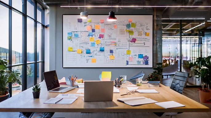 An open office with a large whiteboard covered in sticky notes, diagrams, and flowcharts. Laptops and documents are spread across the desk, symbolizing teamwork and organized project planning.