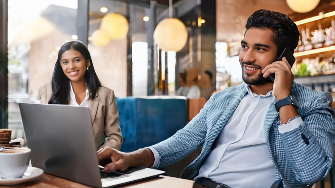 An entrepreneur sits in a coffee shop, multitasking on a laptop and phone, while a personal assistant nearby manages tasks, illustrating effective work-life balance through PA support.