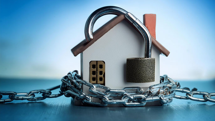 A small model house wrapped in heavy chains with a large padlock, symbolizing the real estate lock-in effect, where homeowners are "locked" into their properties due to financial constraints or market factors.