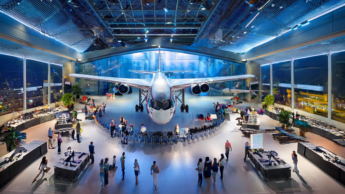 A large exhibition space featuring a modern aircraft at the center, surrounded by attendees exploring aerospace technology displays and interactive booths at a conference.