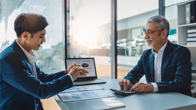 A senior businessman receiving financial advice from a younger professional in a modern office, highlighting retirement planning in Japan's financial sector.