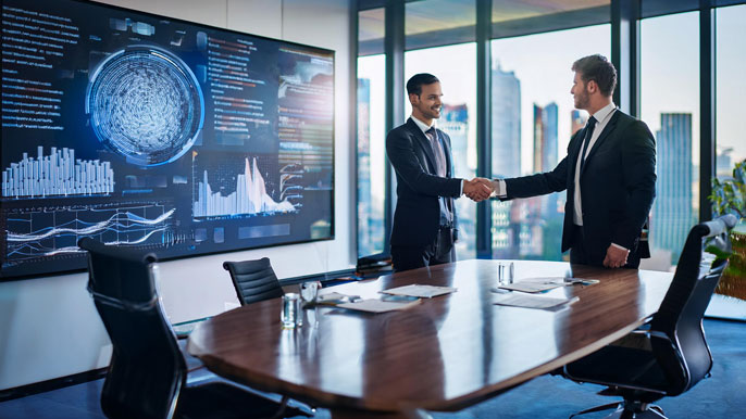 Two business executives shaking hands in a modern conference room, with data charts displayed on a large screen, symbolizing successful business interaction and nonverbal communication.