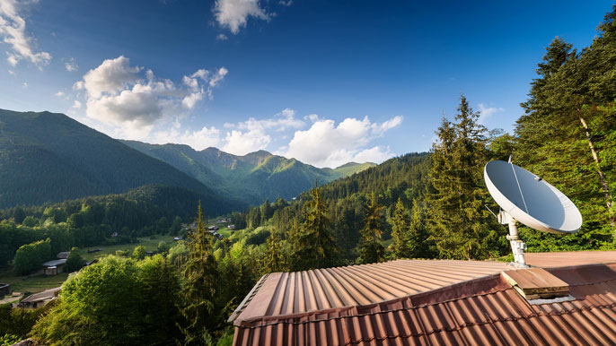 Satellite dish on a rooftop in a remote mountain village, surrounded by lush forests and high peaks, symbolizing connectivity reaching isolated areas.