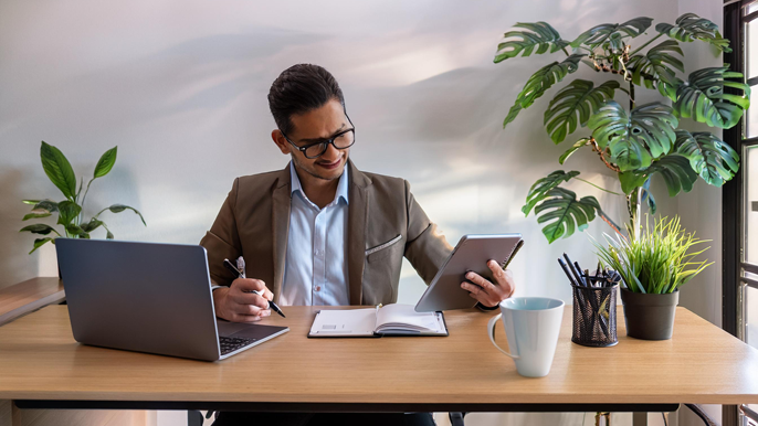 An entrepreneur sitting at a desk with a laptop, tablet, notebook, coffee mug, and indoor plants, representing a productive and well-organized workspace for business success.