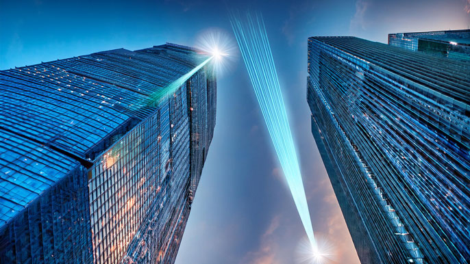 Two skyscrapers connected by visible laser beams between rooftop antennas, illustrating Free Space Optics (FSO) technology providing high-speed data communication across urban buildings without the need for physical cables.