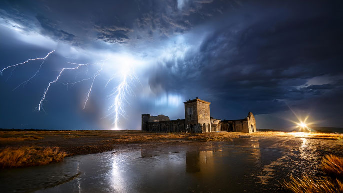 Powerful lightning strike illuminating a dark, stormy sky over an open field, with reflections on the wet ground and an old building in the background, capturing the intensity of the storm.