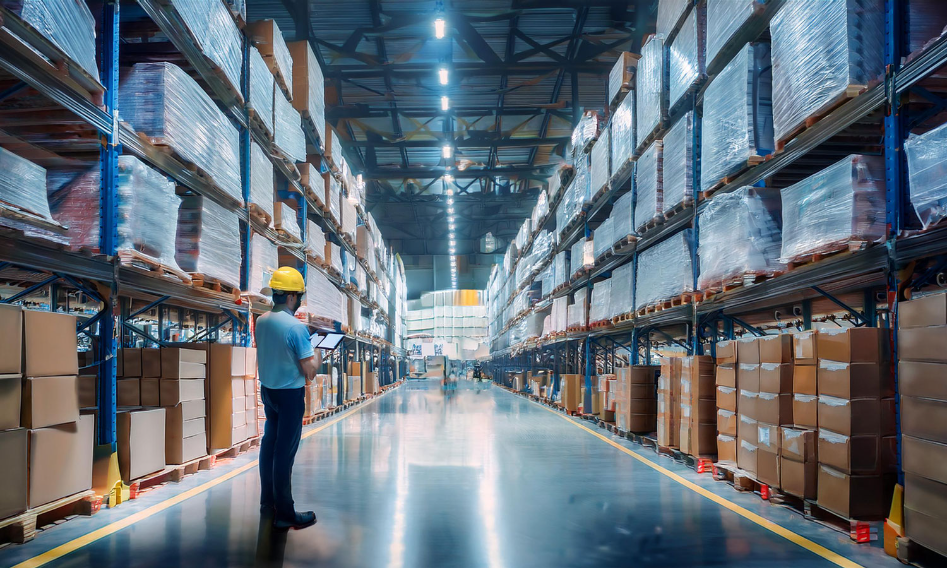 Warehouse worker using a tablet to check inventory in a large, organized warehouse, highlighting stock movement and auditing processes.