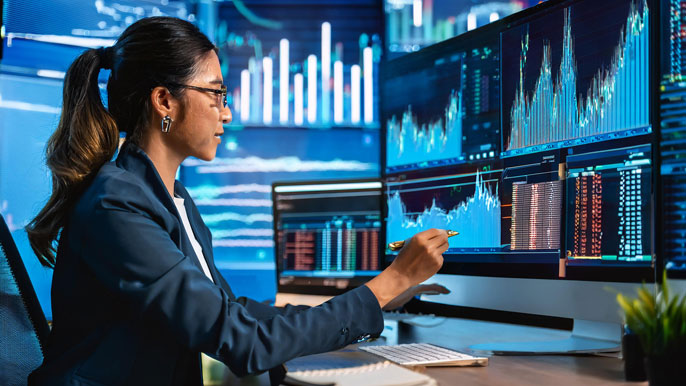 A financial analyst reviewing stock charts and data on multiple computer screens, analyzing ROI and P/E ratios for investment decisions in a modern office setting.