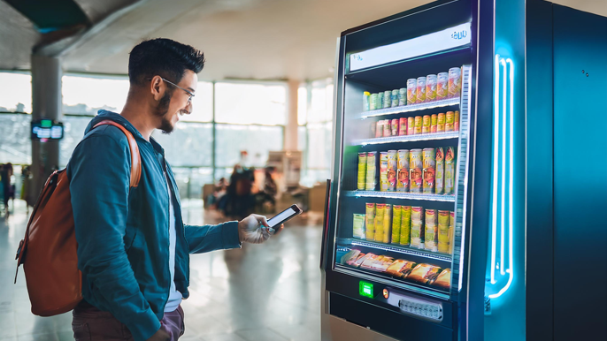 Customer using a mobile wallet for a contactless payment at a sleek, modern smart vending machine offering drinks and snacks in a busy airport setting